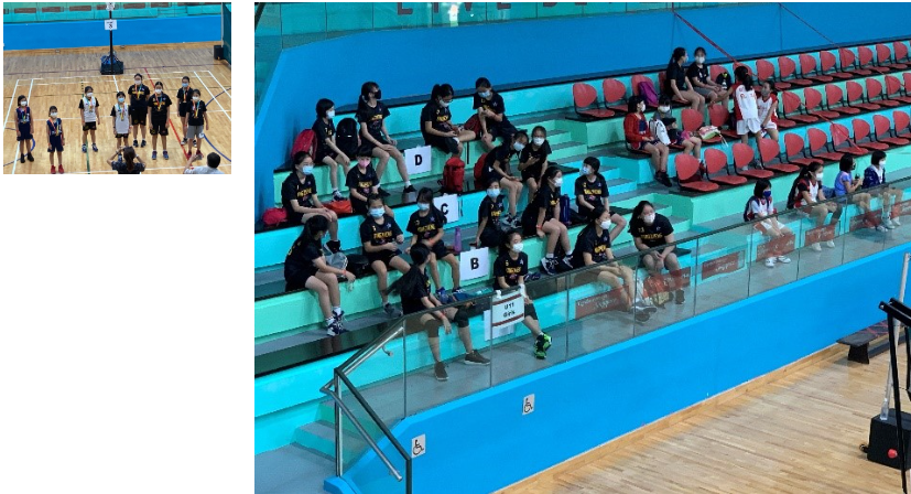 A group of primary school girls basketball players seated at a basketball court watching a basketball match